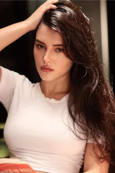 Woman with long dark brown hair wearing a white top, resting her hand on her head and looking at the camera.