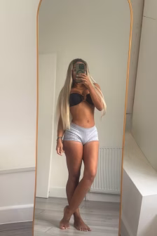 Person taking a mirror selfie in a bright room, wearing a black bra and light gray shorts, with long blonde hair.