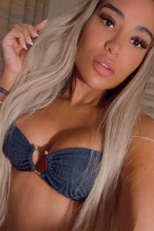 Close-up selfie of a woman with long blonde hair wearing a dark blue bikini top, looking at the camera with slightly parted lips.